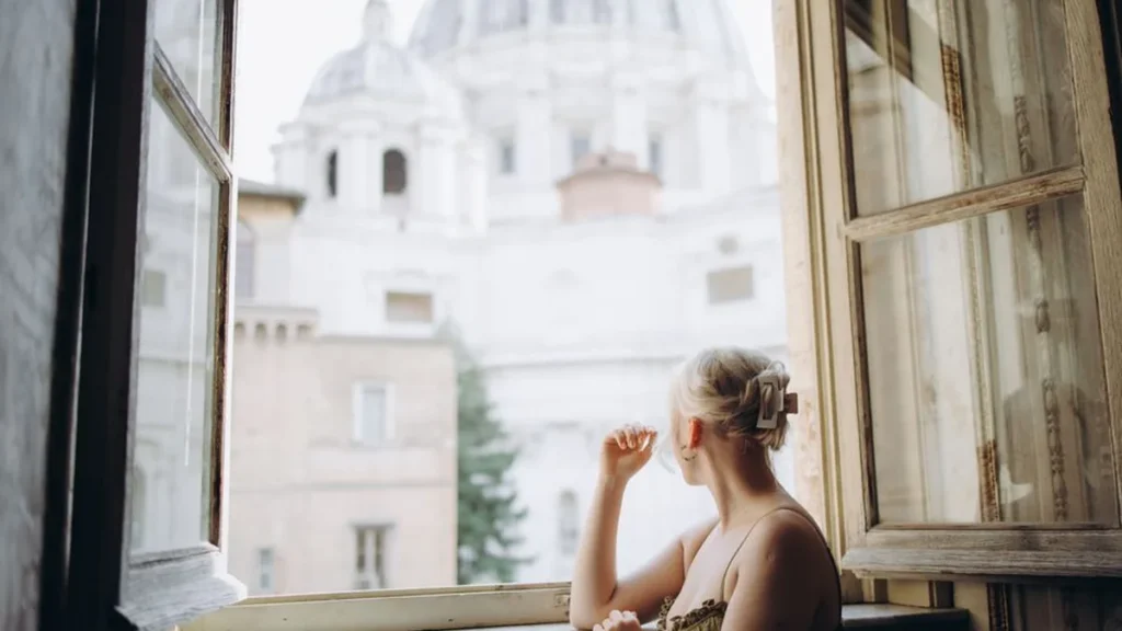 A stylish woman looking out from a beautiful Roman apartment balcony overlooking historic city rooftops.