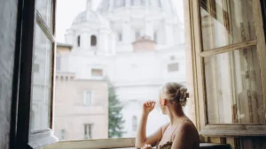 A stylish woman looking out from a beautiful Roman apartment balcony overlooking historic city rooftops.